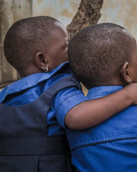 Two small children in blue uniforms, seen from behind, one with an arm round the other